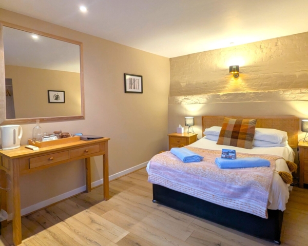 A cozy hotel room with a double bed, wooden headboard, bedside tables with lamps, a wooden desk with a mirror, a tray with a kettle and cups, and neatly folded blue towels on the bed. Warm lighting creates a welcoming atmosphere.