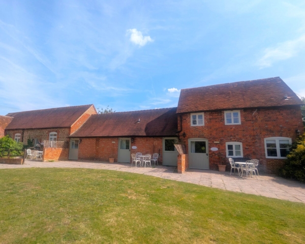 A row of 3 red brick cottages with white-trimmed windows and a sloped roof, set on a stone patio with 3 decorative metal tables and chairs, overlooks a grassy lawn under a blue sky with scattered clouds.