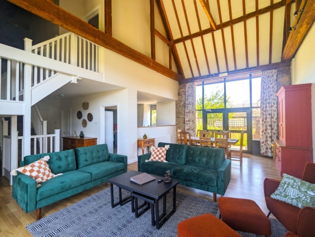 A spacious living room with high vaulted ceilings and exposed wooden beams, featuring green sofas, a patterned rug, a dining area with large windows, and natural light filling the space.