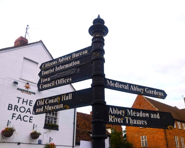 A black signpost with directional signs pointing to local attractions, including Citizens Advice Bureau, Tourist Information, Medieval Abbey Gardens, Abbey Meadow and River Thames, stands in front of brick buildings.