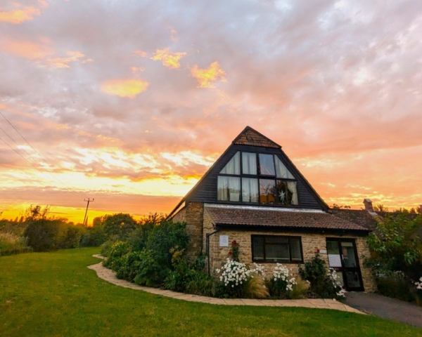 A stone barn with a triangular roof, large windows and reception sign sits among greenery and white flowers. The sky behind is bright with a dramatic orange and yellow sunset, casting a warm light over the scene.